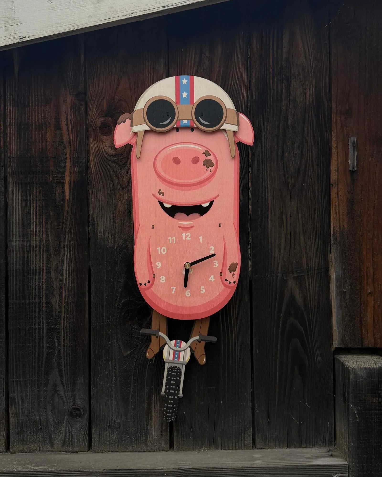 Whimsical pig-shaped wall clock with goggles and a bike, mounted on rustic wood.